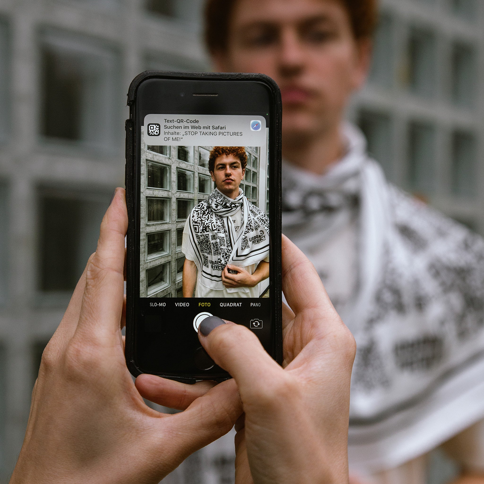 Ein Handy ist auf einen Mann mit einem Schal aus Tencel gerichtet. Auf dem Schal sind viele schwarz weiße QR-Codes in unterschiedlichen Größen zu sehen. Auf dem Bildschirm erscheint ein Text "stop taking pictures of me".