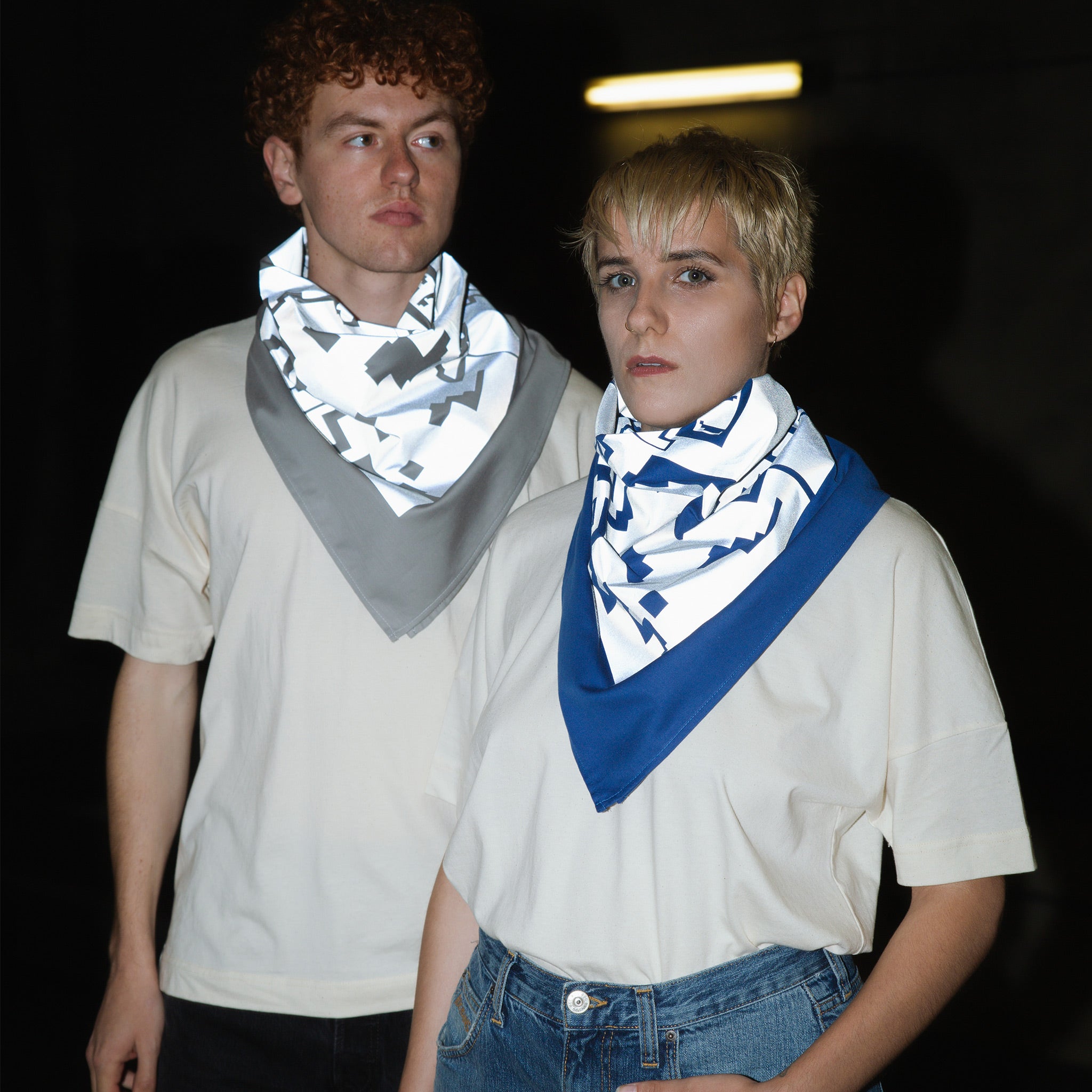 Eine Frau und ein Mann, tragen beide ein großes Bandana in blau und grau um den Hals. Auf dem Bandana ist ein großflächiger Foliendruck in grau zu sehen. Das Foto wurde mit Blitzlicht aufgenommen, es ist die Reflektion durch das Tuch zu sehen.