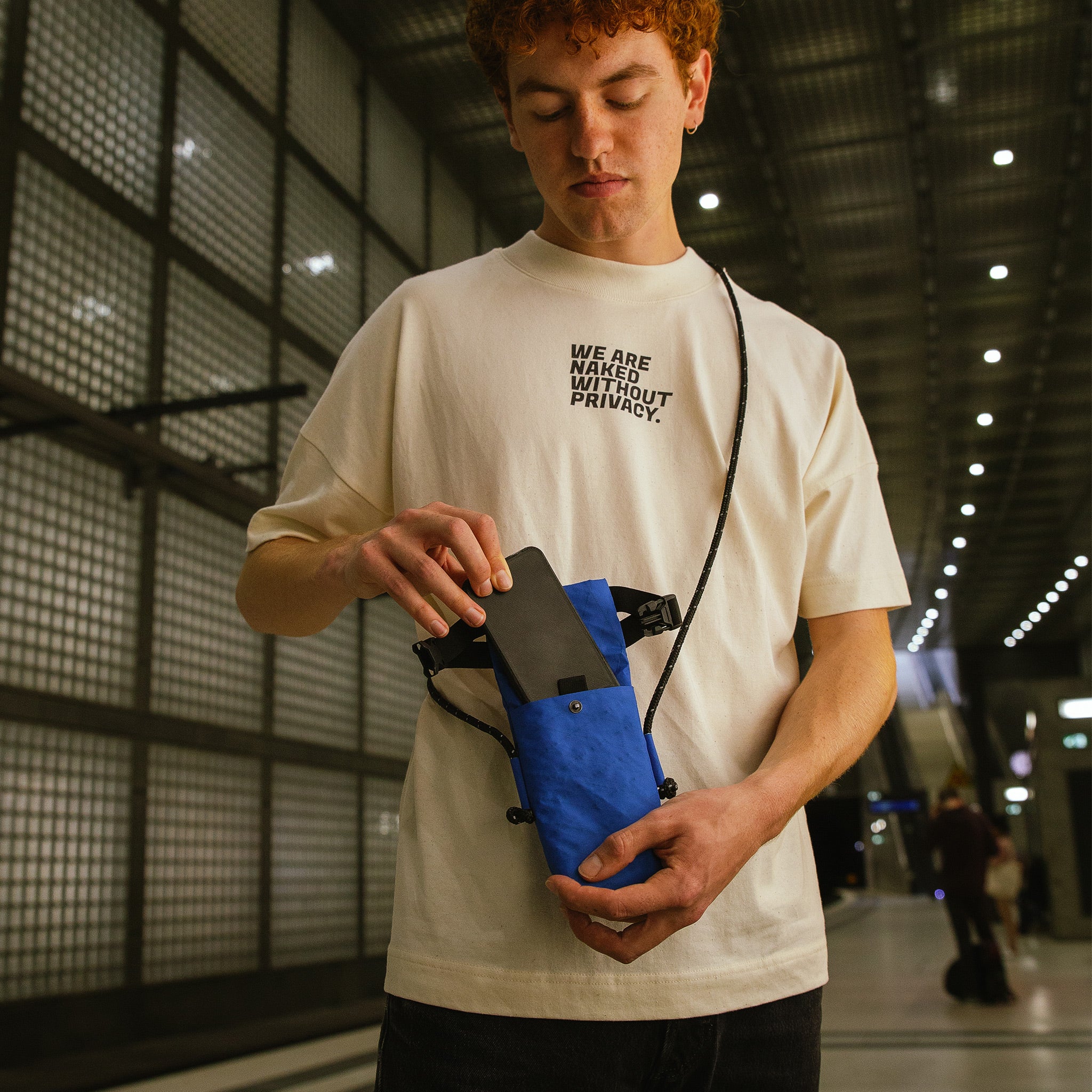 Ein Mann steckt sein Handy in das Onlinefach seiner abschirmenden Umhängetasche von Urbanprivacy. Die Tasche ist blau.