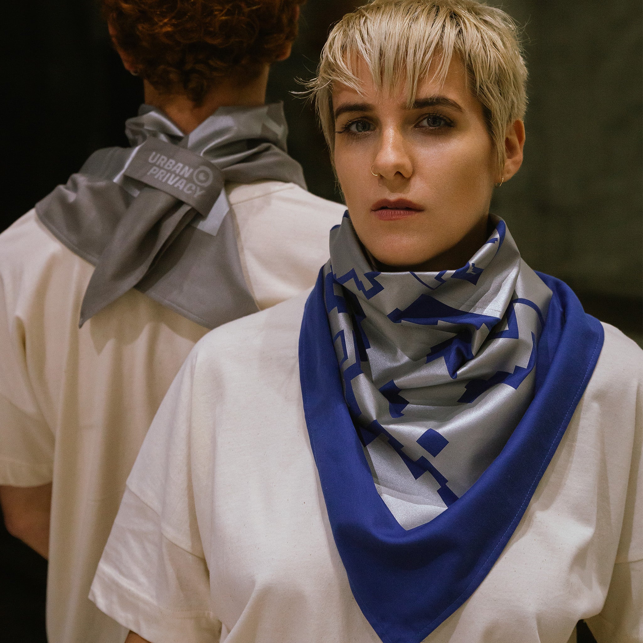 Eine Frau trägt im Vordergrund des Bildes, ein großes Bandana in blau. Auf dem Bandana ist ein großflächiger Foliendruck in grau zu sehen um den Hals.