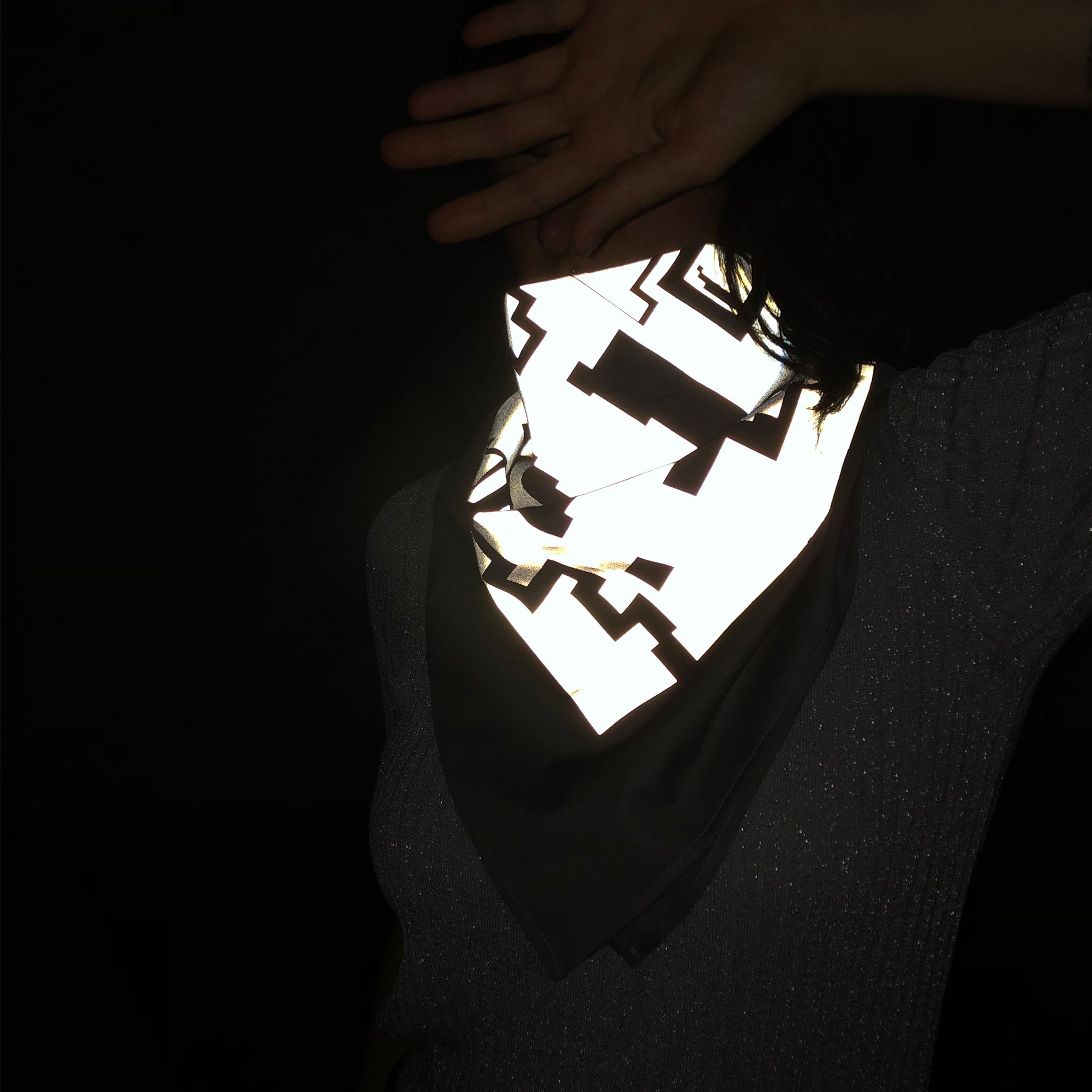 Eine Frau trägt ein großes Bandana um den Hals. Das Foto wurde in einer dunklen Umgebung mit Blitzlicht aufgenommen, es ist die Reflektion durch das Tuch zu sehen.